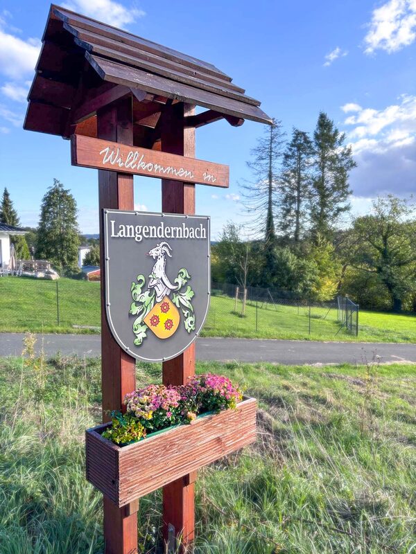 Überdachtes Holzschild „Willkommen in Langendernbach“ mit dem Ortswappen – Ziegenbock über einem Schild mit drei Rosen – und einem bepflanzten Blumenkasten.